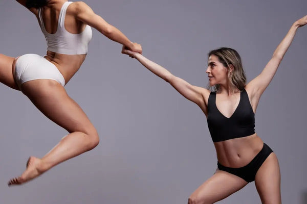 Two models in dynamic poses wearing a white bralette and second-skin thong set and a black racerback bra and brief set against a grey backdrop.